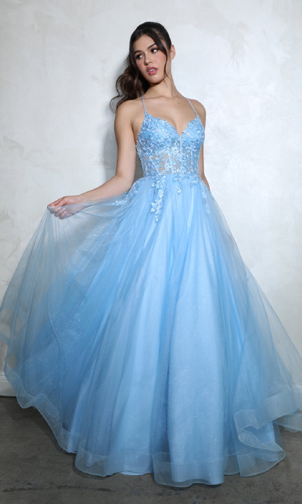 A woman stands against a plain light wall wearing the Lace-Up Corset Long Prom Ball Gown: Eureka 9960, a light blue, floor-length A-line dress featuring a sheer bodice, floral appliques, and thin straps.